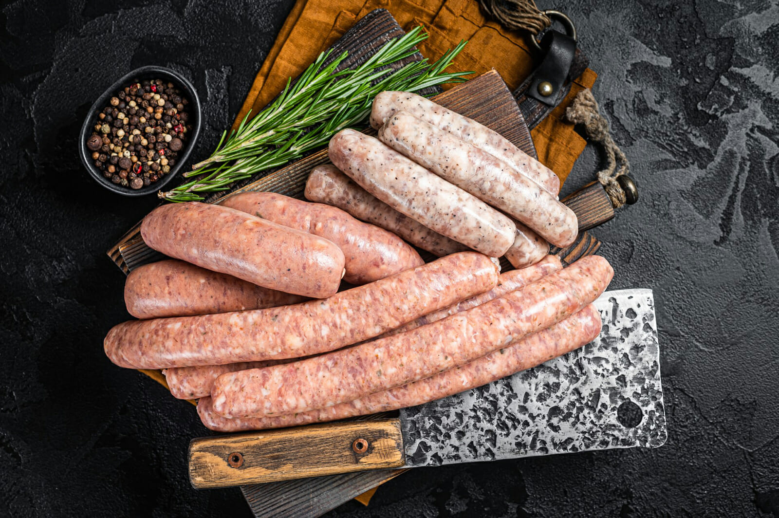 Assorted Raw Sausages On A Wooden Board With Rosemary And Peppercorns.